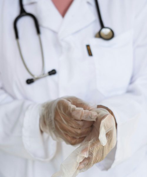 Faceless ethnic medical worker in lab coat and stethoscope taking of transparent gloves after approaching patients for examination in modern hospital
